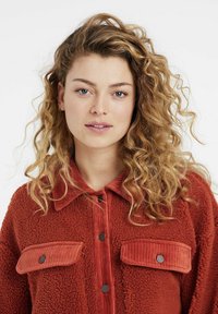 Rust-colored jacket made from plush textured fabric, featuring a corduroy collar and patch pockets with black buttons.