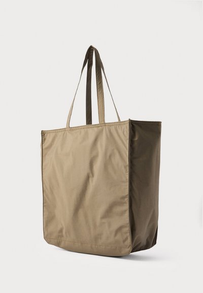 Sac fourre-tout rectangulaire beige avec deux longues anses, en tissu lisse, debout sur un fond clair uni.