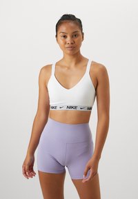 Hvit sports-bh med svart Nike-logo bånd, kombinert med høy midje, lyse lilla shorts. Glatt tekstur og tettsittende design.