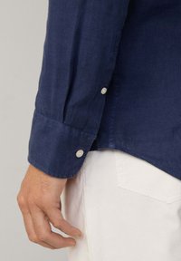 Navy blue long-sleeve shirt with a buttoned cuff, made from a textured fabric. White button visible, with a curved hemline and light-colored pants.