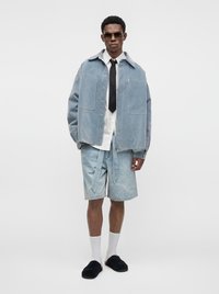 Jeune homme portant une veste bleu clair oversize, une chemise rayée, une cravate noire, un short en jean, des chaussettes blanches, des chaussures noires sans lacets et des lunettes de soleil.
