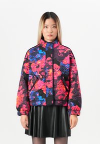 Woman wearing a puffy floral jacket in bright pink, blue, and black, paired with a black leather skirt and sheer black tights.