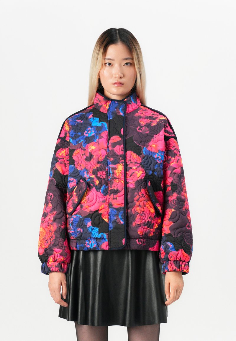 Woman wearing a puffy floral jacket in bright pink, blue, and black, paired with a black leather skirt and sheer black tights.
