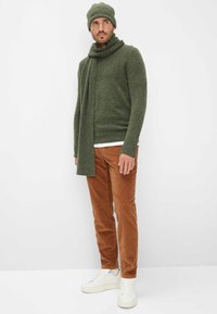 Maglione verde scuro a trama con berretto e sciarpa abbinati, abbinato a pantaloni in velluto a coste color ruggine e scarpe da ginnastica bianche.