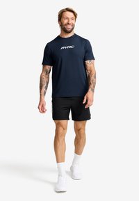 Marinblå atletisk t-shirt med "RVRC"-logotyp, kombinerad med svarta shorts. Modellen bär vita sneakers och synliga mönstrade tatueringar på armarna.