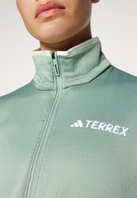 Svijetlozelena Terrex jakna na zip s visokim ovratnikom, s bijelim izvezenim logotipom na prsima i glatkim, teksturiranim materijalom.