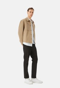 Veste beige à boutons avec deux poches poitrine, associée à une chemise blanche, un pantalon noir et des baskets blanches, sur fond uni.