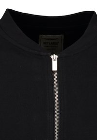 Chaqueta negra con cremallera central plateada y cuello redondo, mostrando la etiqueta "KEY LARGO hecho a mano desde 1994" en el interior del cuello.
