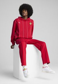Rød hættetrøje og matchende bukser med hvide lodrette striber, der har et Ferrari-logo. Hvide sneakers med røde accenter og logo.