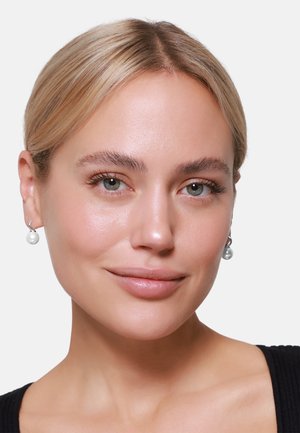 Silberne Creolen mit einer einzelnen hängenden Perle. Das Model hat blonde Haare, einen glatten Teint und trägt ein schwarzes Oberteil.