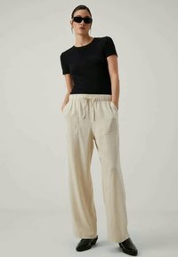 T-shirt noir ajusté à col rond, pantalon large crème avec taille élastique et poches, associé à des lunettes de soleil noires et des bottes à talons.