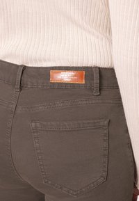 Jeans in denim verde oliva con etichetta in pelle marrone, design a cinque tasche e dettagli di cucitura visibili. Parte superiore a coste beige parzialmente visibile.