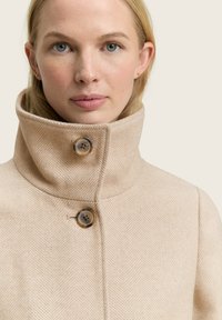 Vrouw met lichte huid en blauwe ogen draagt een beige herringbone hoge-kragen jas, vastgezet met twee grote knopen.