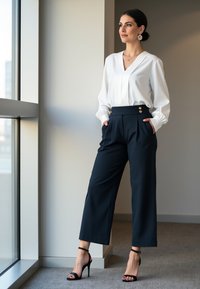 Biała bluzka z bufiastymi rękawami, granatowe spodnie z wysokim stanem z złotymi guzikami i czarne sandały na obcasie, stojąca przy oknie.