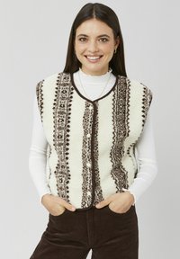 Veste en polaire crème avec détails et boutons à motifs marron, dotée d'un col rond et d'une coupe sans manches. Associée à une chemise blanche à manches longues.