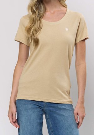 Kvinde iført beige t-shirts med korte ærmer og lille hvidt logo på brystet samt blå jeans, stående mod en ensfarvet lys baggrund.