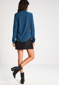 Blusa lunga color teal con colletto, abbinata a una mini gonna in suede nera e stivali alla caviglia neri. Texture liscia, design slim fit.