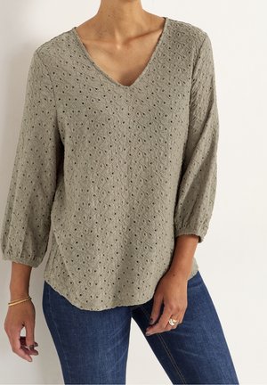 Donna che indossa una camicetta verde oliva chiaro testurizzata con maniche a tre quarti e jeans blu, mostrando la parte superiore del corpo e le braccia.