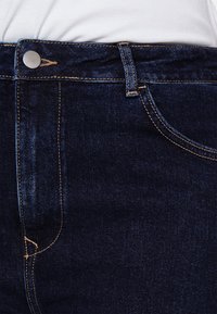 Tmavě modré džíny z denimu se hladkým povrchem, s pruhovaným stříbrným knoflíkem, kontrastním žlutým prošíváním a předními kapsami.