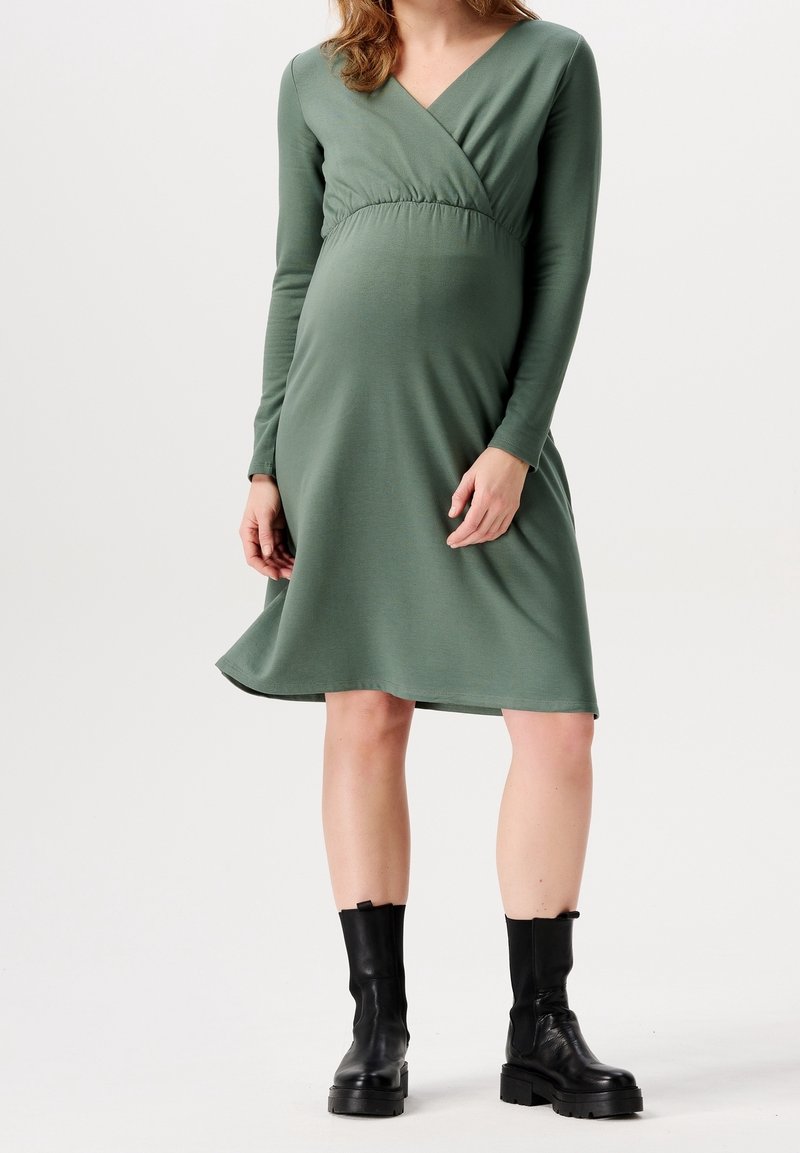 Femme enceinte portant une robe verte à manches longues, longueur genou, et des bottines noires, debout devant un fond uni.