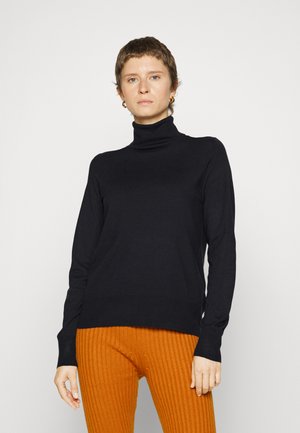 Pull à col roulé noir en tissu doux avec des manches longues et une silhouette ajustée. Porté avec un pantalon orange côtelé.