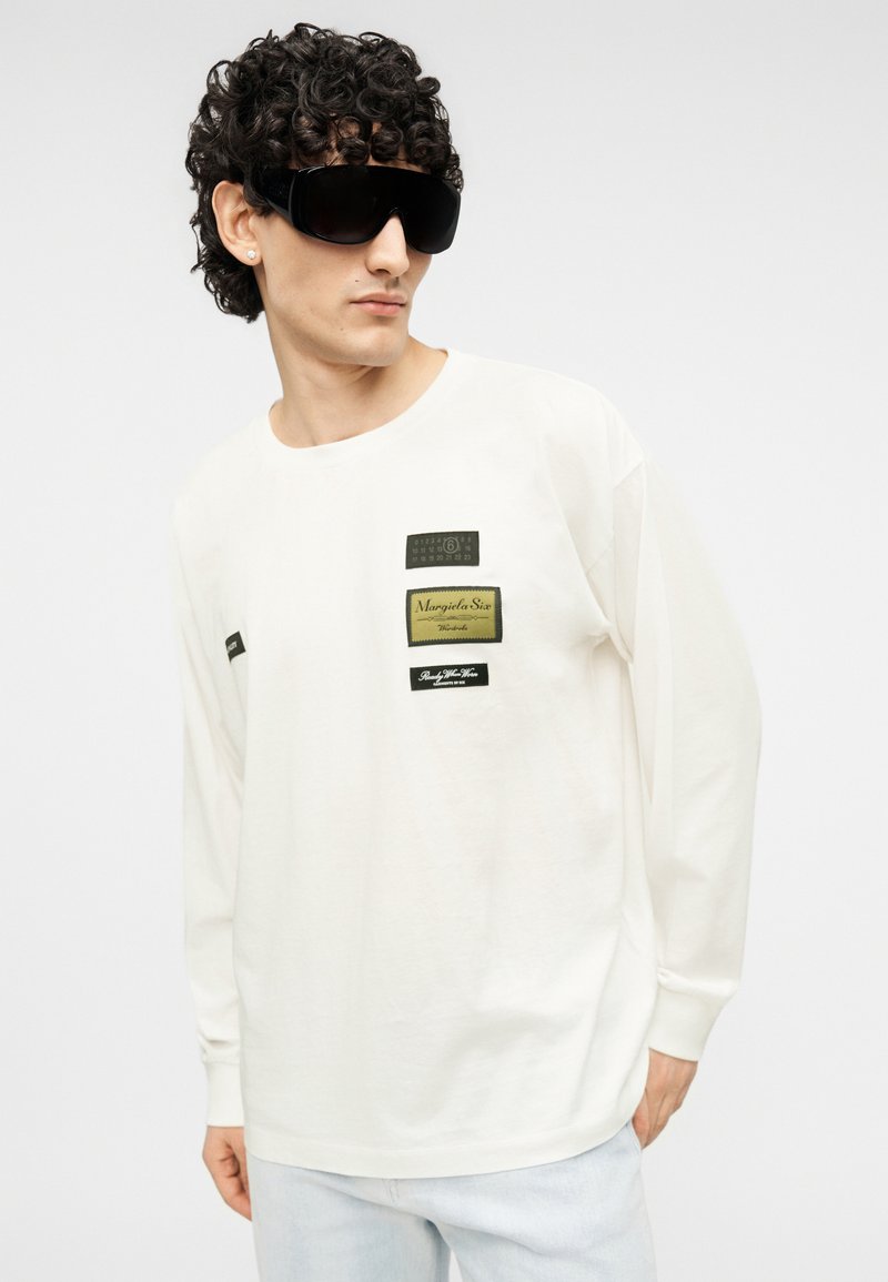 Maglietta bianca a maniche lunghe con molteplici etichette del marchio nere e verdi sul petto. Il modello indossa occhiali da sole neri oversize.