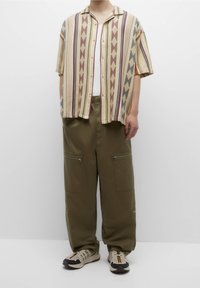 Camicia a maniche corte con bottoni, decorata da motivi a zigzag e strisce multicolori; abbinata a pantaloni cargo larghi di colore verde oliva con tasche con zip.