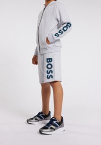 Šedá mikina na zip s bočními kapsami a nápisem "BOSS" na rukávu, doplněná o odpovídající šortky s nápisem "BOSS" výraznými písmeny. Tenisky s modrými akcenty.