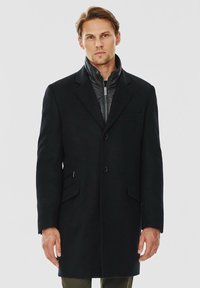 Czarny długi płaszcz o dopasowanym kroju, z stójką, dwoma przednimi kieszeniami i zapięciem na guziki. W środku znajduje się pikowana czarna warstwa.