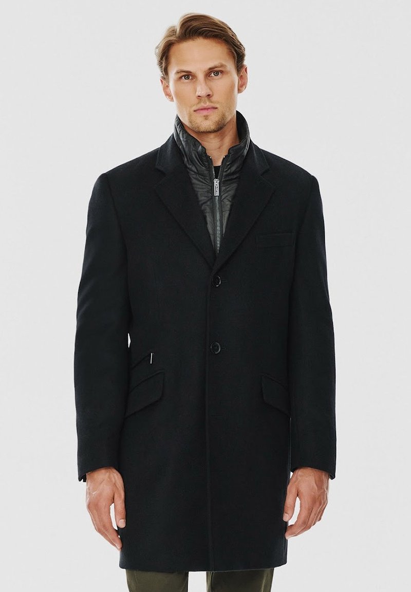 Czarny długi płaszcz o dopasowanym kroju, z stójką, dwoma przednimi kieszeniami i zapięciem na guziki. W środku znajduje się pikowana czarna warstwa.
