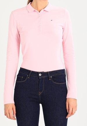 Mujer vestida con una camisa polo de manga larga ajustada de color rosa claro y jeans azul oscuro, de pie contra un fondo blanco liso.