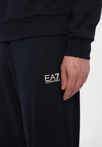 Meresinine spordipüksid pehme tekstuuriga, elastse vöökoha ja vasakul jalal oleva valge logoga. Suumvaade vöökoht ja käe interaktsioon.