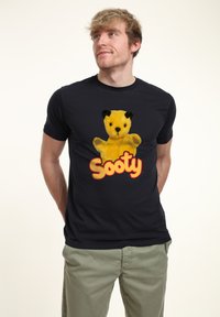 Svart t-shirt med en illustration av en gul björn med svarta öron, tillsammans med texten "Sooty" i orange och gula bokstäver.