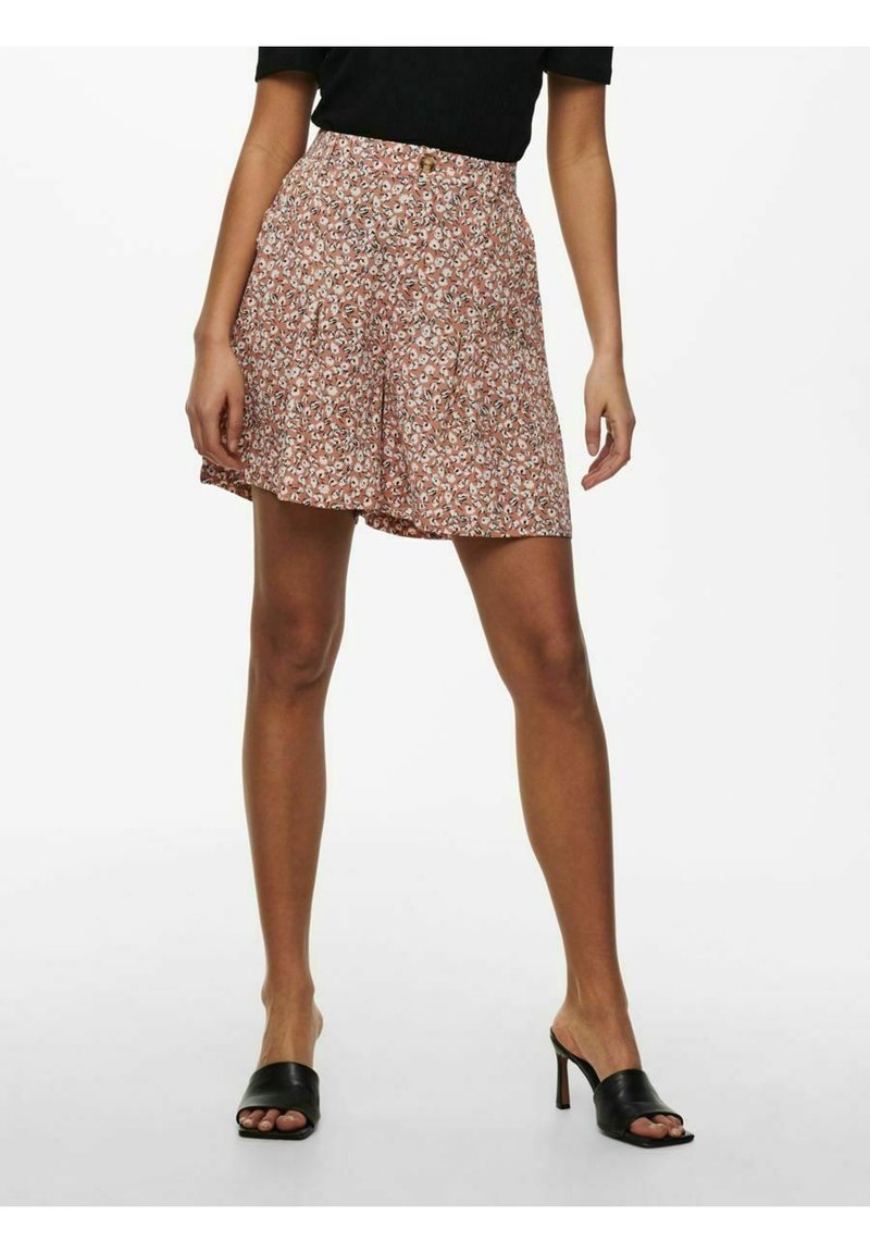 Femme portant un short à motifs floraux roses associé à des mules à talons noires et un haut noir, debout devant un fond blanc.