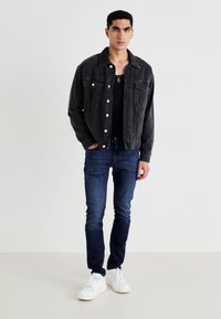 Chaqueta de mezclilla negra con cierre de botón, dos bolsillos en el pecho, combinada con jeans azul oscuro y zapatillas blancas, de pie contra un fondo liso.