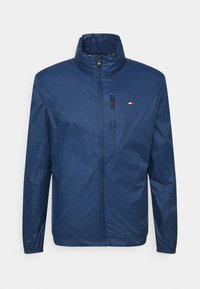 Chaqueta ligera azul marino con cremallera frontal, cuello alto y puños elásticos. Cuenta con un pequeño logo en el pecho y un bolsillo con cremallera.