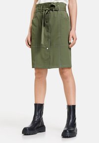 Olivgrüne Rock mit Frontknöpfen und Gürtel, ausgestattet mit seitlichen Taschen und kontrastierenden weißen Nähten, getragen mit schwarzen klobigen Stiefeln.