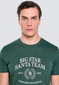 Zelené tričko s krátkým rukávem s bílým grafickým textem "BIG STAR SANTA TEAM" se středovým designem vavřínu a logem. Vyrobeno z měkkého bavlněného materiálu.