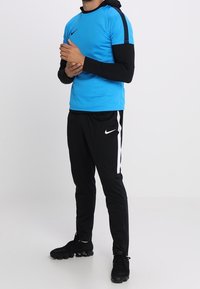 Niebieska koszulka sportowa z krótkim rękawem i czarnymi akcentami, czarne spodnie z białymi paskami oraz czarne buty sportowe. Materiał wydaje się lekki i elastyczny.