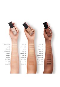 Three hands holding foundation bottles next to swatches on arms. Bottles have black caps; swatches show a range of skin tone shades.