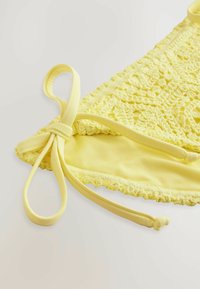 Kollane heegeldatud bikiiniseelik ühtlase mustriga, seotud rihmadega ja siledast voodrist, mille allääres on laineline äär.