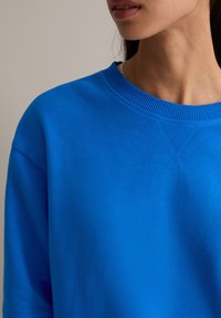 Niebieska bluza z okrągłym dekoltem, ściągaczem przy kołnierzu i teksturowaną tkaniną. Charakteryzuje się widocznym przeszyciem i luźnym krojem.