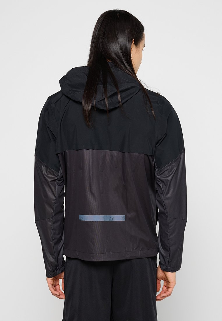 Persoon met lang, donker haar, gekleed in een twee-kleurige zwarte waterdichte jas met capuchon, weergegeven van achteren tegen een effen achtergrond.