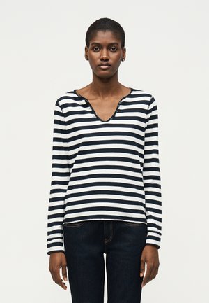 Femme debout devant un fond blanc, portant une chemise à rayures noires et blanches à manches longues et un jean foncé, regardant droit devant.