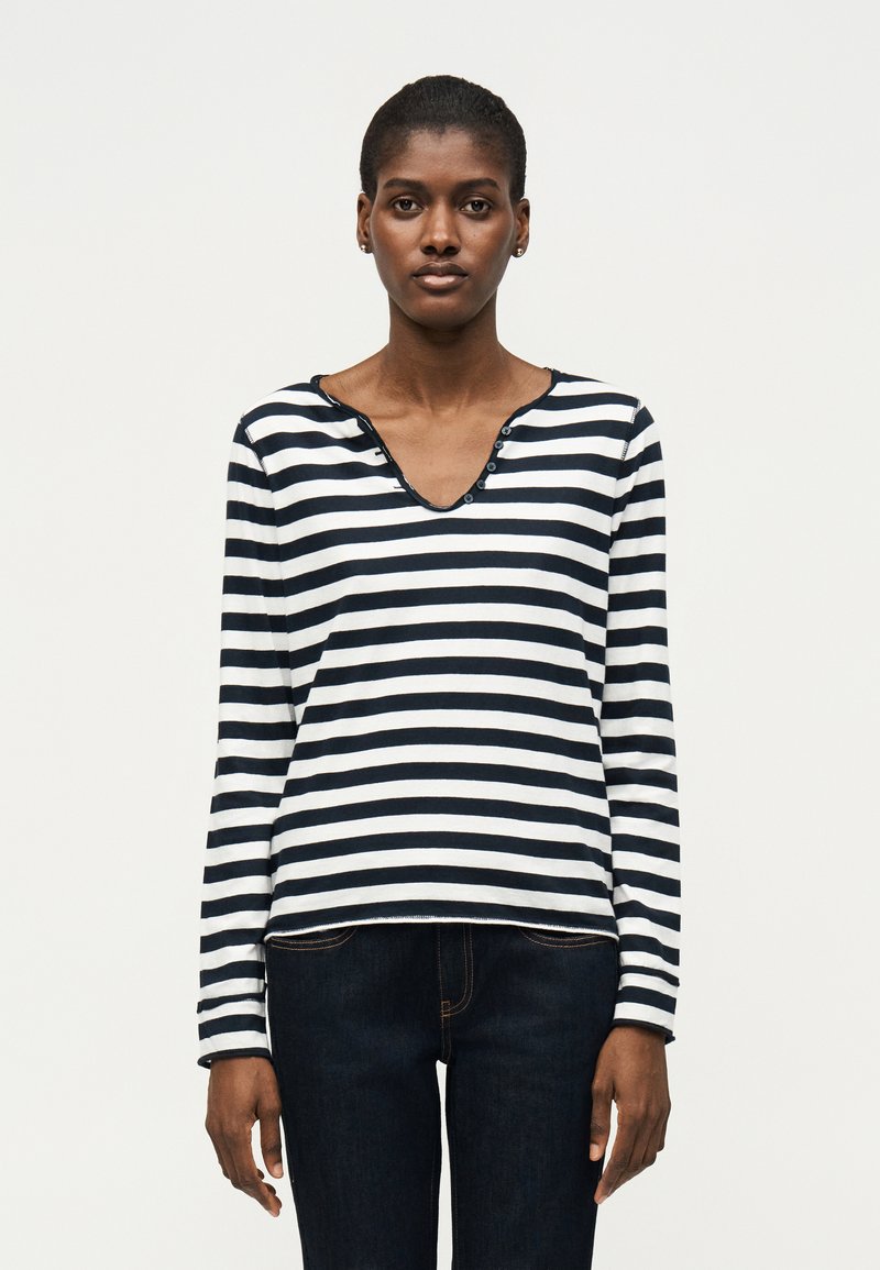 Γυναίκα που στέκεται μπροστά σε λευκό φόντο, φοράει μια μακρυμάνικη μαύρη και άσπρη ριγέ μπλούζα και σκούρο τζιν, κοιτάζοντας ευθεία μπροστά.