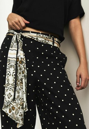 Person trägt schwarze Hosen mit weißen Punkten und einen weiß-braun gemusterten Schal als Gürtel, kombiniert mit einem schwarzen kurzen Oberteil.