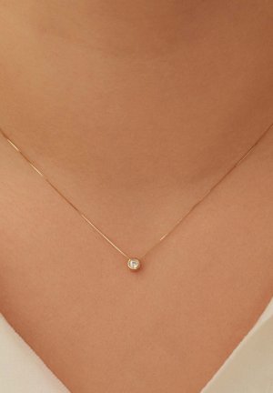 Collier délicat en chaîne d'or avec un petit pendentif rond serti d'un diamant, reposant sur le cou d'une personne au-dessus d'un haut blanc.