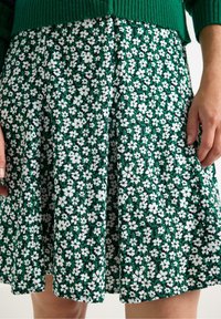 Grøn blomsterkjole med et tæt mønster af hvide blomster, plisseret design, blød tekstur og mid-length snit. Stof ser let og luftigt ud.