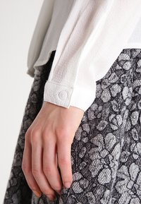 Haut blanc texturé à manches longues avec un poignet à boutons, porté sur une jupe grise à motifs comprenant des designs floraux et lacés.
