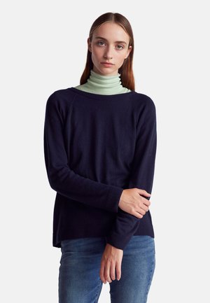 Maglione lungo blu navy in materiale morbido con scollatura rotonda. Indossato sopra un dolcevita verde chiaro, abbinato a jeans blu.